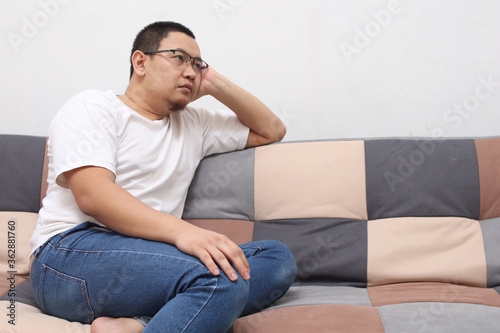 Unhappy Asian man sitting and thinking on his sofa at home in the living room, sad expression