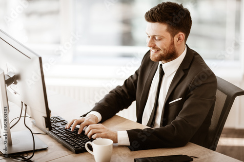 Business succsess concept. Happy smiling young businessman sitting in office and working on computer. Man in suit indoors on glass window background photo