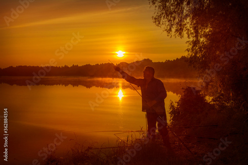 Morning on the lake dawn. A fisherman is standing on the shore next to fishing rods.