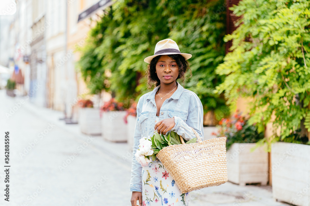 Beautiful afro american girl in straw sun hat, holding wicker bag with bouquet of peonies walking vintage area with european view in summer bright day. Travel, student, relax, lifestyle concept.