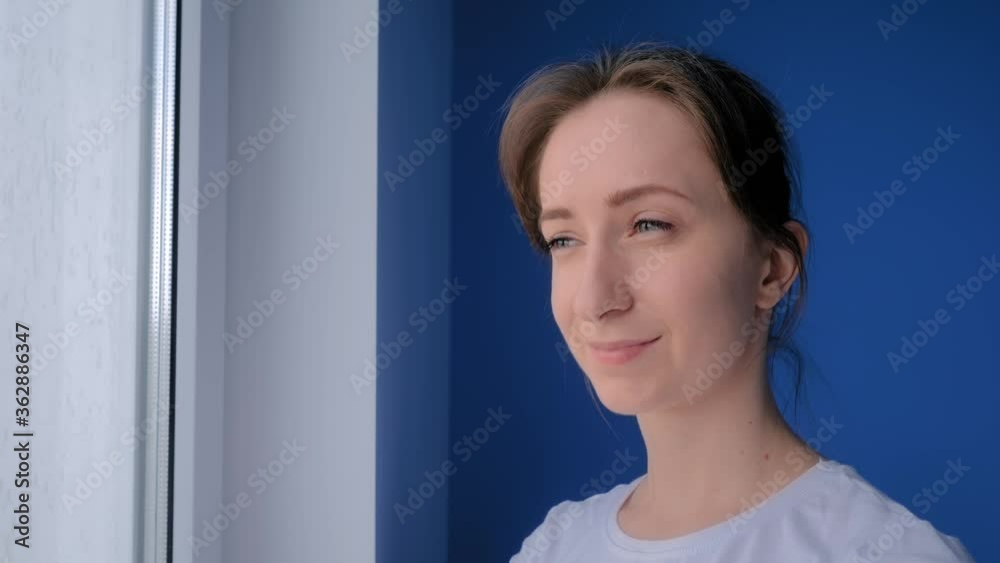 Slow motion: portrait of young positive woman in white shirt smiling and looking out of window in room with blue wall at home. Self isolation, prevention, quarantine, leisure time, safety concept Slow motion: portrait of young positive woman in white shirt smiling and looking out of window in room with blue wall at home. Self isolation, prevention, quarantine, leisure time, safety concept