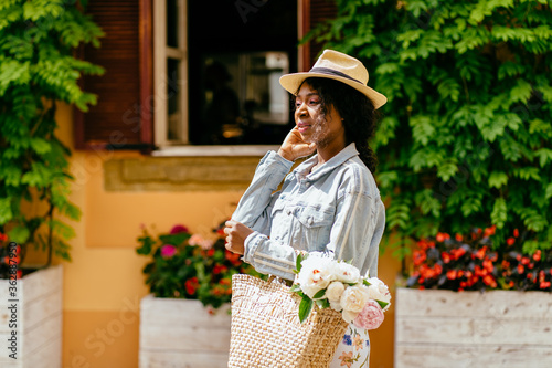 Beautiful african american woman with curly hair wearing fashionable sunglasses, holding smart phone in hand, walking on the italian street at sunny summer day.