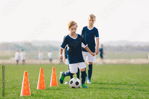 Boys training unit on soccer field. Young footballers practice dribbling skills on the grass pitch. Kids running between red soccer training cones. Summer soccer camp for children team
