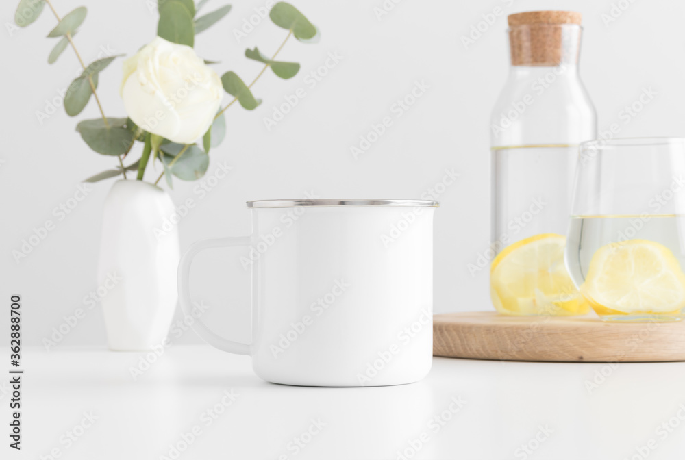 Enamel mug mockup with flowers in a vase, glass and a bottle on a white table. Enamel mug mockup with flowers in a vase, glass and a bottle on a white table.