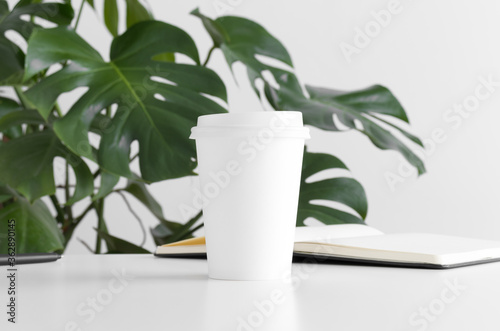 Coffee paper cup mockup  with a monstera plant and a notebook on a white table.