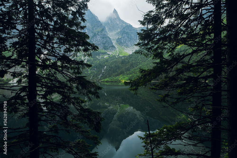 Beautiful view of foggy mountains cover by dark clouds and green forest with a reflection in a lake. Stony shore. Morskie Oko. Marine Eye. High Tatras, Zakopane