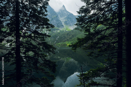 Beautiful view of foggy mountains cover by dark clouds and green forest with a reflection in a lake. Stony shore. Morskie Oko. Marine Eye. High Tatras, Zakopane