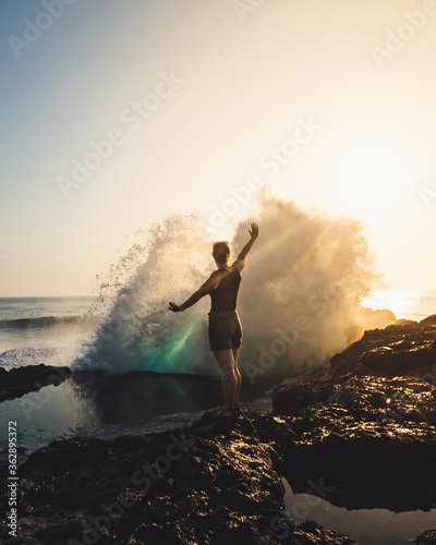 Silhouette of a young woman standing on a cliff side with her arms raised and waves crashing in