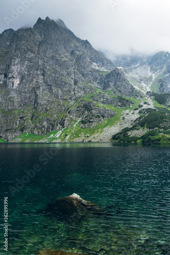 Scenic view of foggy mountains cover by dark clouds and green forest with a reflection in a lake. Stony shore. Morskie Oko. Marine Eye. High Tatras, Zakopane