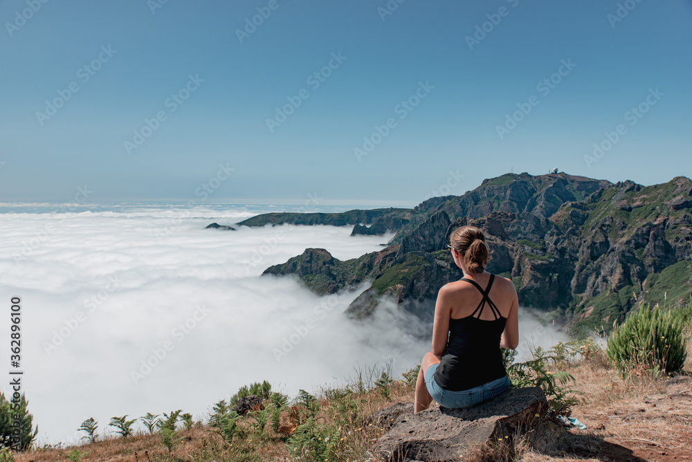 Young brunette woman with sunglasses sitting at the edge of the mountain over the clouds