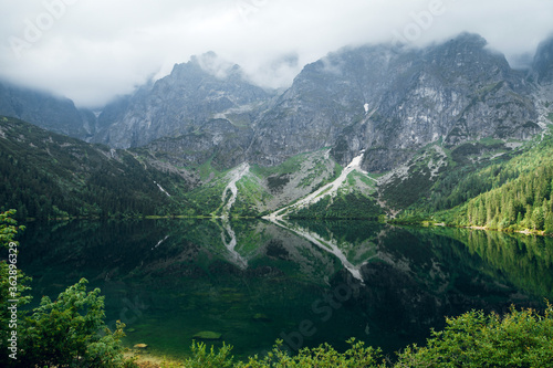 Beautiful view of foggy mountains cover by dark clouds and green forest with a reflection in a lake. Stony shore. Morskie Oko. Marine Eye. High Tatras, Zakopane