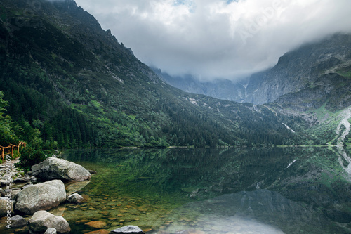 Beautiful view of foggy mountains cover by dark clouds and green forest with a reflection in a lake. Stony shore. Morskie Oko. Marine Eye. High Tatras, Zakopane