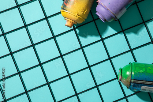 Top view of used spray paint cans on top of a metal grid, on cyan background with free space for text