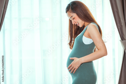 20 weeks pregnant woman in a gray dress, posing comfortably in her bedroom By standing at the edge of the window curtain and in a happy smile
