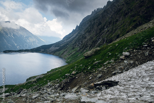 Beautiful view of foggy mountains cover by dark clouds and green forest with a reflection in a lake. Stony shore. Morskie Oko. Marine Eye. High Tatras, Zakopane