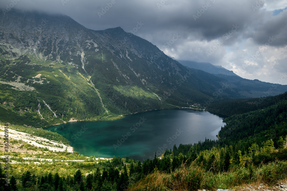 Fototapeta premium Aerial view shot of beautiful green hills and mountains in dark clouds and reflection on the lake Morskie Oko lake, High Tatras, Zakopane