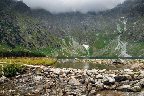 Scenic view of foggy mountains cover by dark clouds and green forest with a reflection in a lake. Stony shore. Morskie Oko. High Tatras, Zakopane