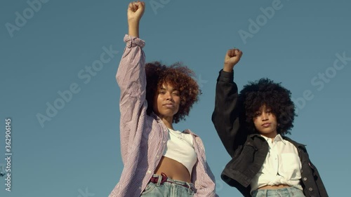 portrait of two mixed-race women with huge afro hair makes a protest black raised fist gesture outdoors portrait on the sky background
