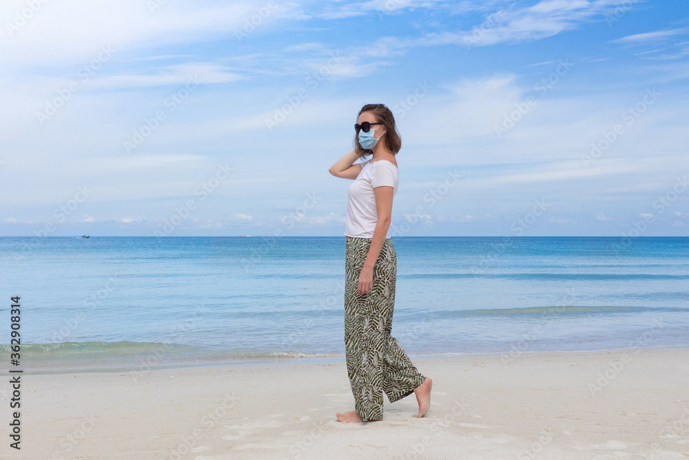 Attractive young woman in black sun glasses and medical mask walking along a tropical beach with sea scape background. Virus prevention concept. New normal.