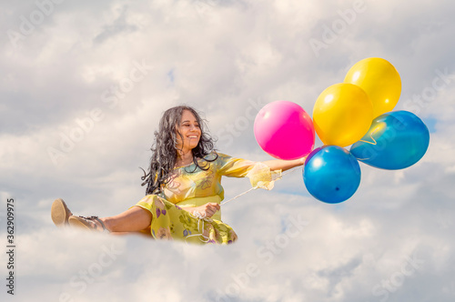Young smiling brunette girl sitting on a cloud and holding colorful balloons in her hands