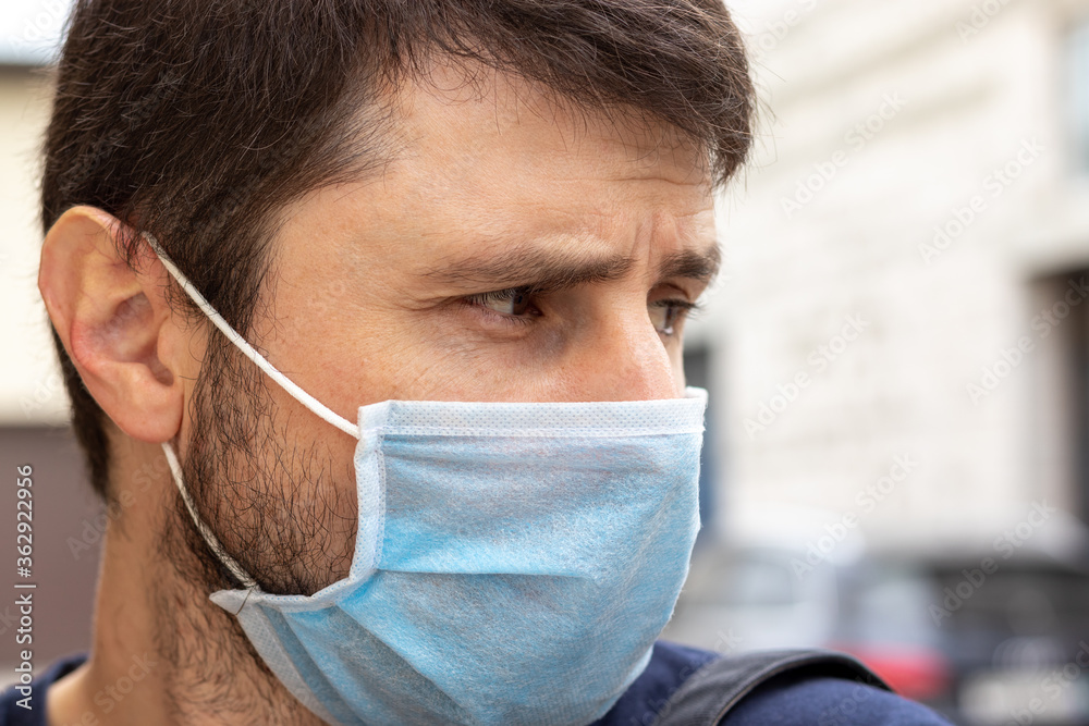 Street closeup portrait of a frowning caucasian man in protective blue medical mask in period of pandemic coronavirus covid-19. Male in a disposable respirator to protect against infection with virus