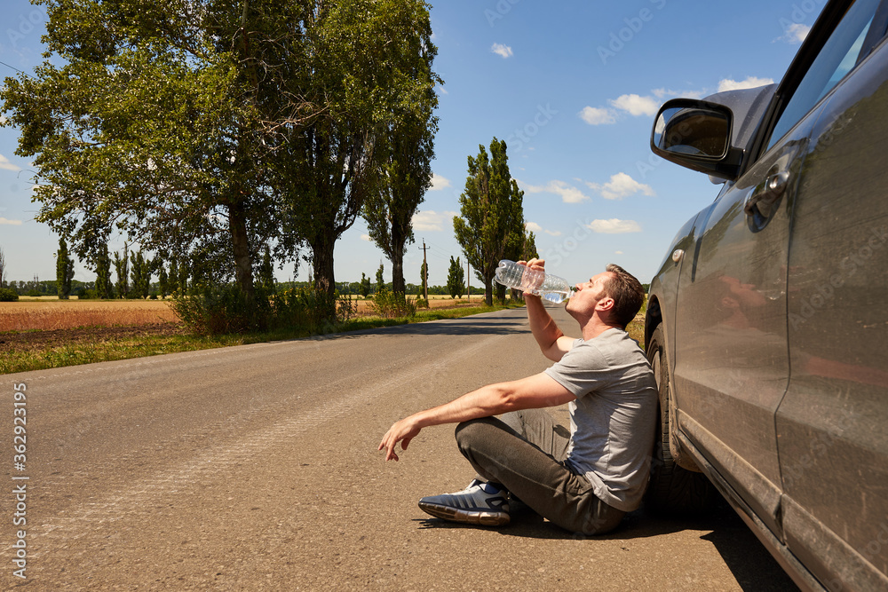 The driver drinks water while sitting on the road near a car with an open hood on a hot sunny day.
