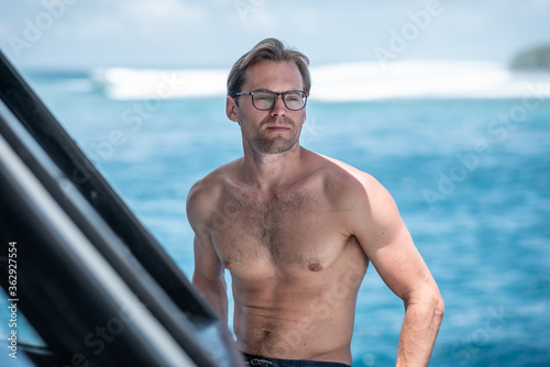 Portrait of handsome shirtless man, surfer in stylish glasses on surfsafari boat and blue ocean with waves behind © Lila Koan
