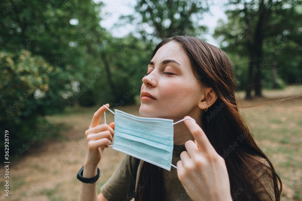 Young caucasian pretty woman removes a medical protective mask from her face on nature, surrounded by trees, breathing clean fresh air Young caucasian pretty woman removes a medical protective mask from her face on nature, surrounded by trees, breathing clean fresh air