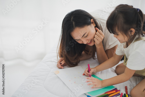 Love of young Asian mother and daughter lie on white bed and enjoy to play creative education drawing in bed room at home which smiling and felling happy. Asian love family live at home concept.