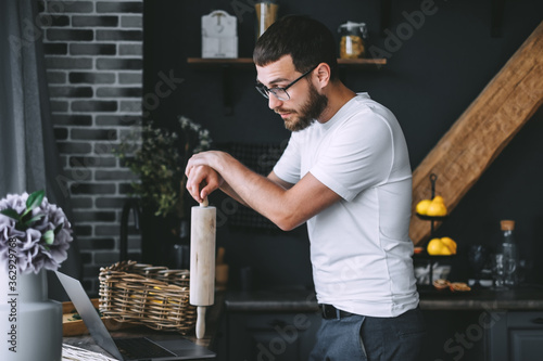 Young pensive caucasian bearded man is looking at food recipes in a laptop, while standing in the kitchen, holding a rolling pin in hands. Cooking preparation.
