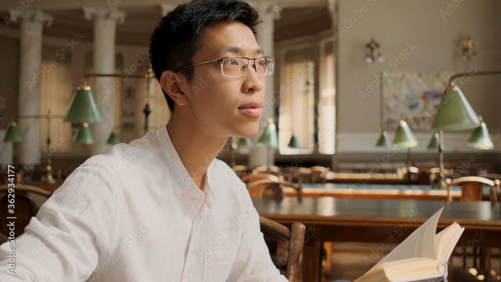 Portrait of asian student with textbook dreamily looking away in library. Young man studying in university