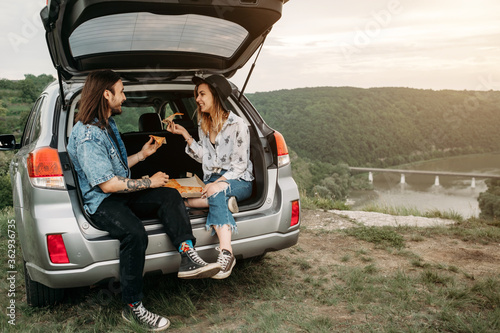 Young Stylish Traveling Hipsters Having Fun Sitting in Car Trunk and Eating Pizza, Travel and Road Trip Concept © Romvy