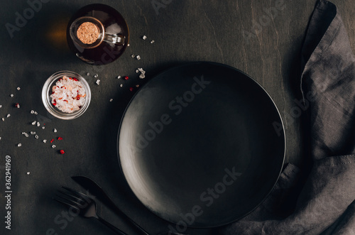 Black table setting: plate, napkin, silverware, olive oil and salt on black wooden background. Top view. Flat lay.