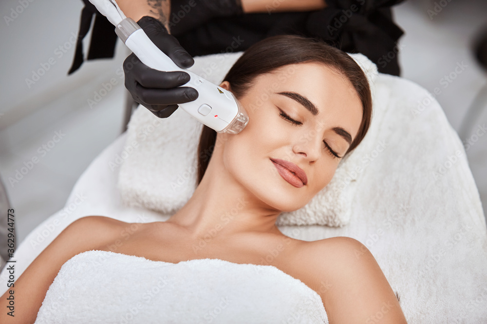 beautiful girl lying in spa salon covering breast with white towel and getting massage for her cheek with professional apparat