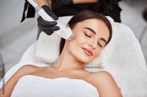 beautiful girl lying in spa salon covering breast with white towel and getting massage for her cheek with professional apparat