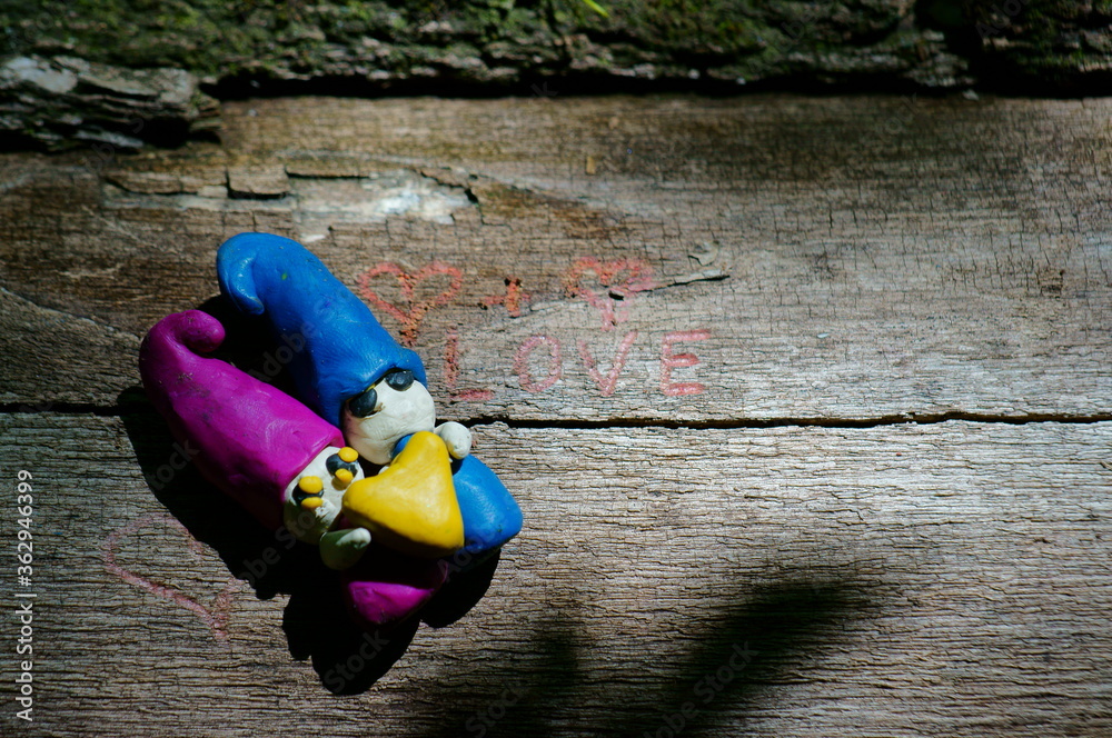Two gnome out of clay holding a heart. Fairy figures on a wooden background. Drawn heart and the inscription love.