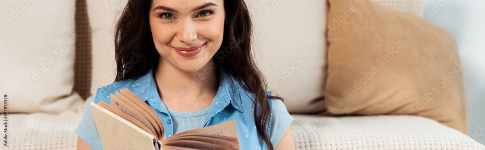 Panoramic shot of smiling woman looking at camera while reading book at home Panoramic shot of smiling woman looking at camera while reading book at home