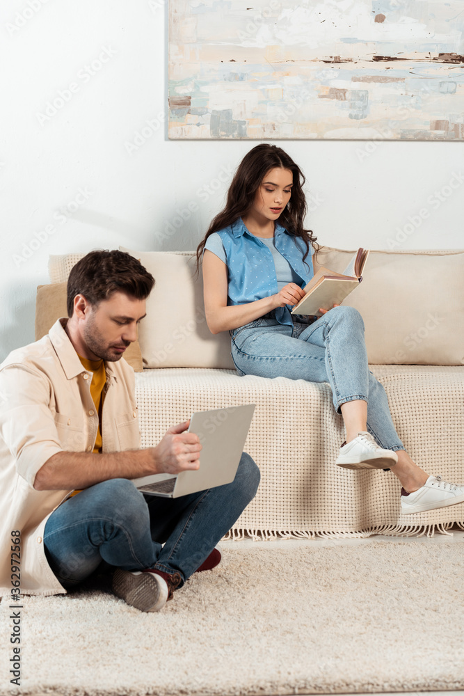 Man using laptop near woman reading book in living room Man using laptop near woman reading book in living room