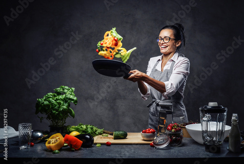 Happy middle-aged female chef tossing chopped vegetables in the air from a frying pan. Healthy food concept. Studio photo on a dark background photo