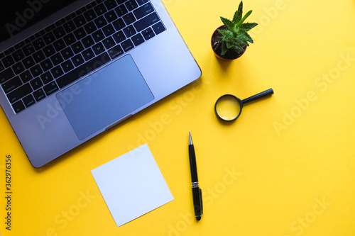 Working space with laptop, plant, magnifying glass, pen and blank sheet on bright yellow background with copy space. Top view, flat lay. Office desk working space. Business and education concept