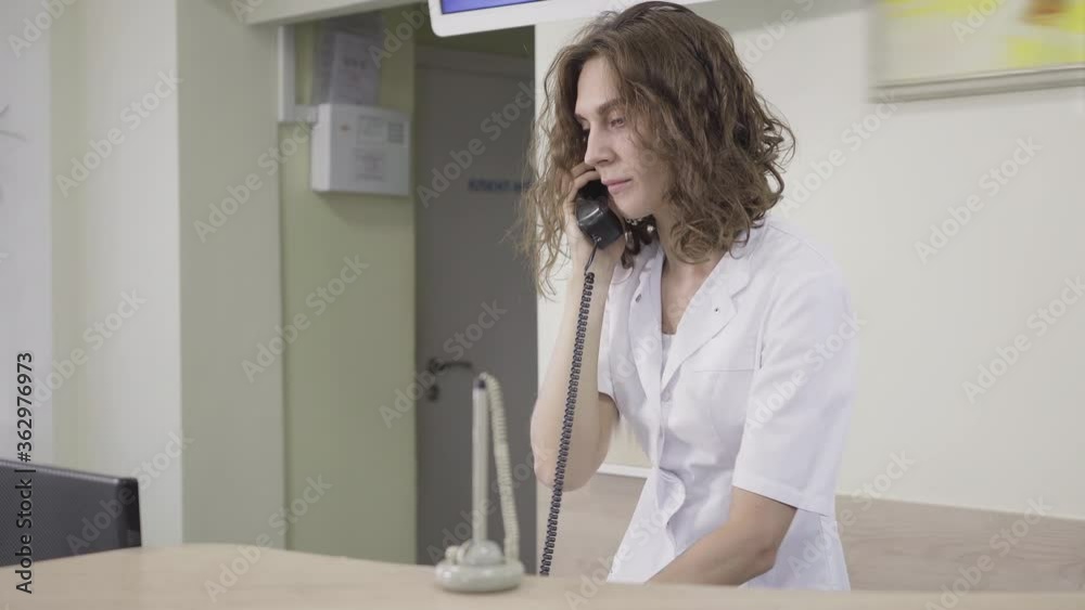 Receptionist receiving call in hospital. Portrait of concentrated Caucasian woman consulting patient. Brunette curly-haired female employee talking on the phone at reception. Medicine, service. Receptionist receiving call in hospital. Portrait of concentrated Caucasian woman consulting patient. Brunette curly-haired female employee talking on the phone at reception. Medicine, service.