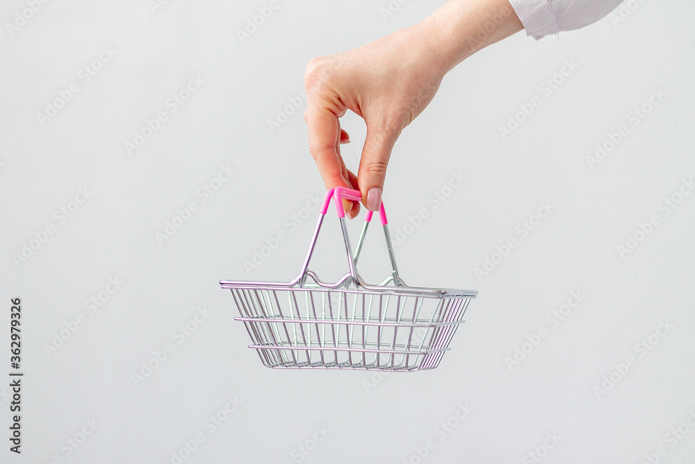 Fototapeta premium Close-up of a woman's hand horizontally holding a toy small metal shopping basket with a pink plastic handle isolated on a white background. copy space for advertising. The concept of shopping.