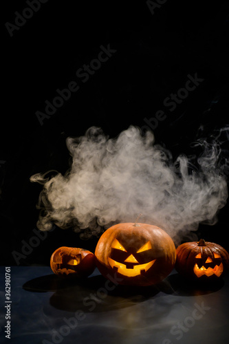 Three creepy halloween steaming pumpkins with a carved luminous smirk on a black background. A handmade jack-o-lantern head with a candle inside in the dark among the fog. Trick or treat. Postcard.