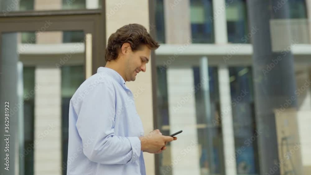 Side view medium tracking shot of young man in casual clothes text messaging on cell phone while walking down city street in slow motion and smiling