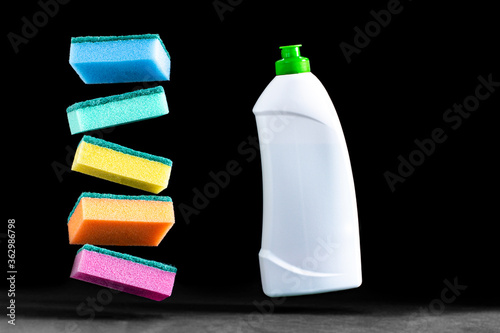dish washing sponges in different colors and a white bottle of cleaning agent levitate in the air above the dark blurred surface of the table