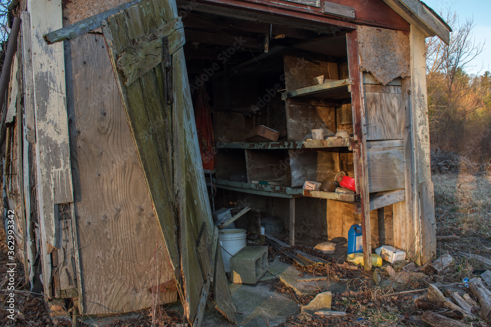 Entrance to an abandoned shed with door hanging off and strewn household items and garden chemicals amidst the debris in rural New England.