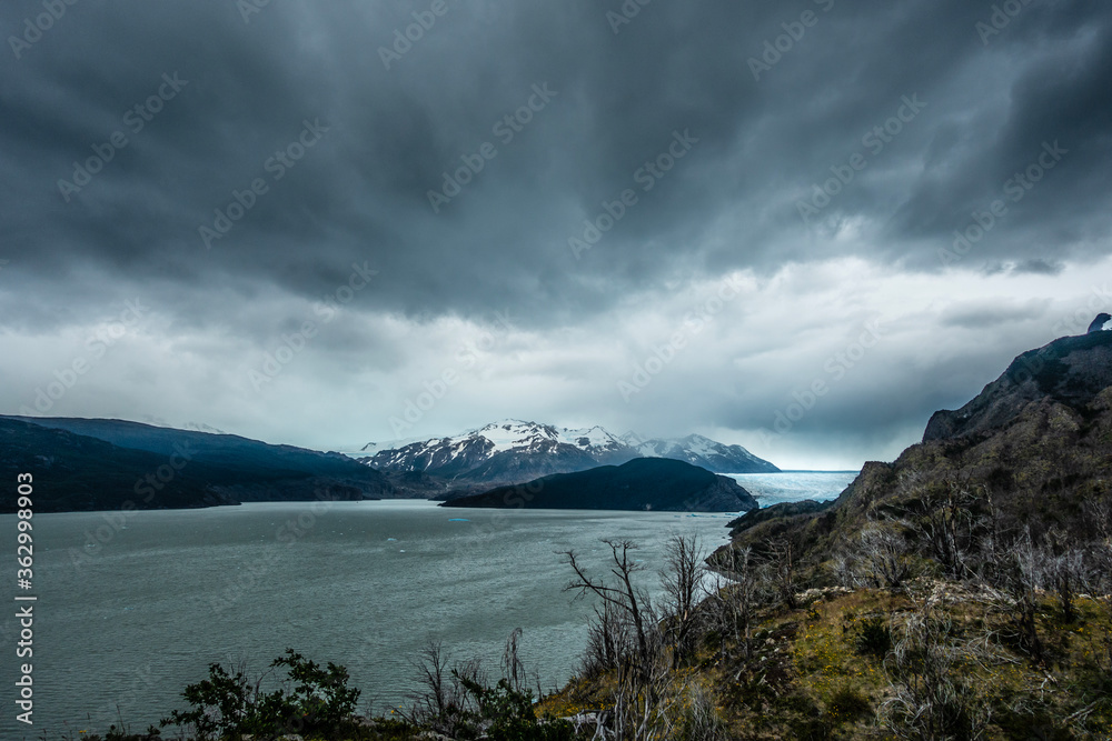 A lake and glacier are seen with snowy mountains and ominous, gloomy clouds. Trees and other foliage as well as rocks appear in the foreground. Torres del Paine National Park, Patagonia, Chile
