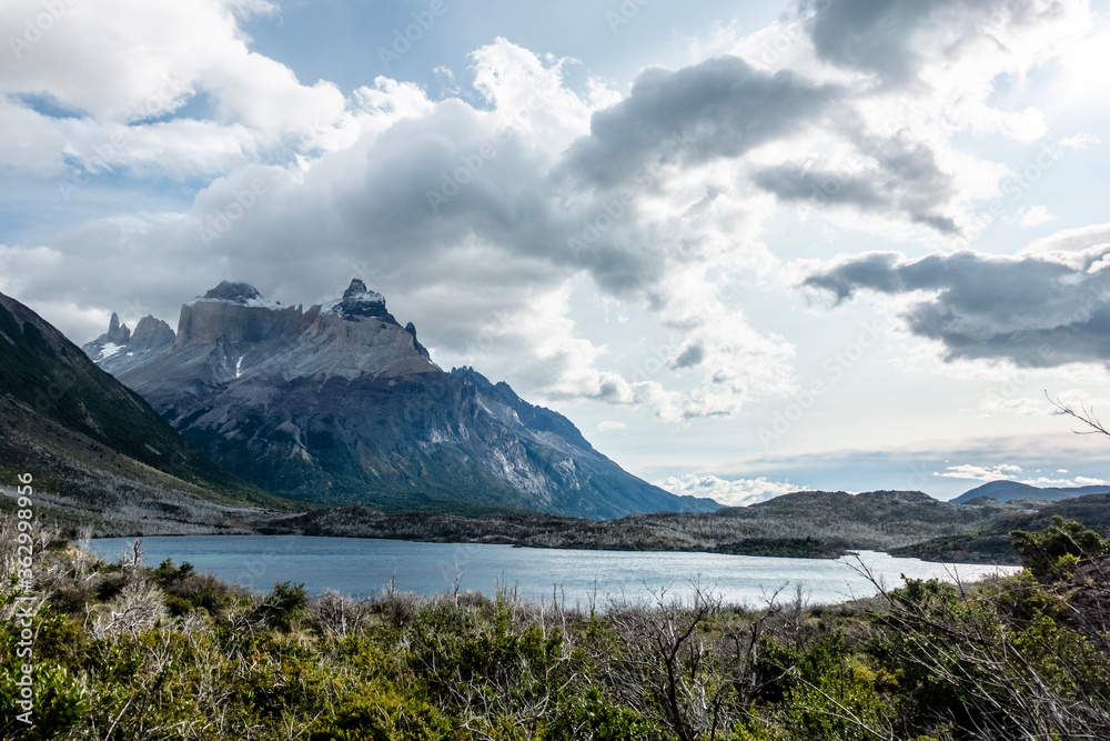 A mountain is seen above a lake with trees in the foreground and clouds above. Some of the trees were previously burnt in a forest fire. Paine Massif, Torres del Paine National Park, Patagonia, Chile