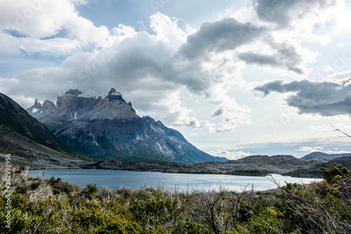 A mountain is seen above a lake with trees in the foreground and clouds above. Some of the trees were previously burnt in a forest fire. Paine Massif, Torres del Paine National Park, Patagonia, Chile