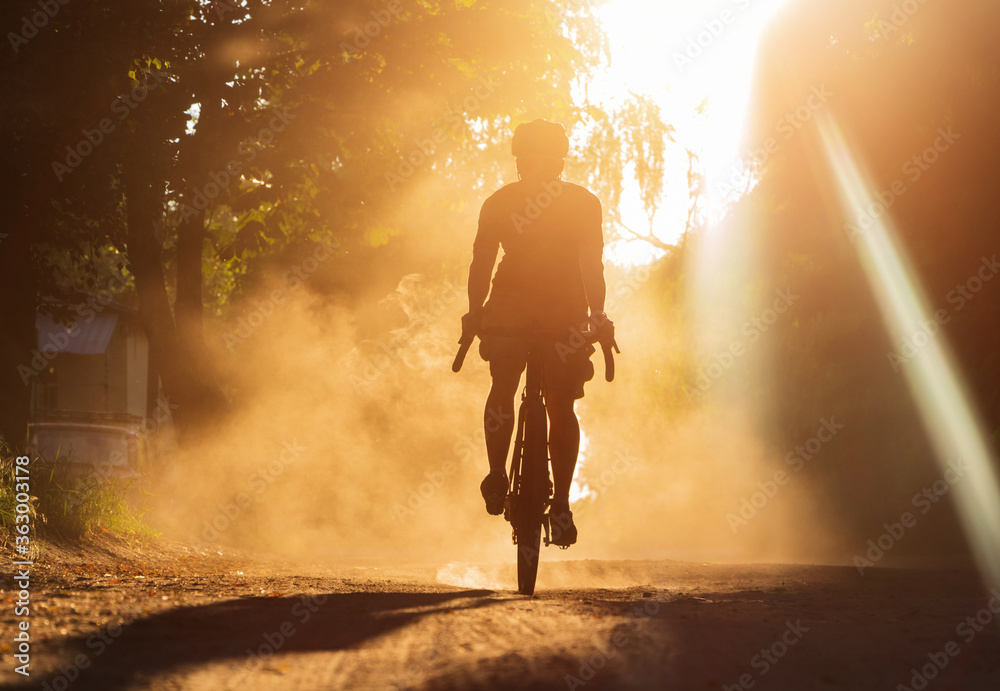 Fototapeta premium A man riding a bicycle on a gravel road at sunset. A silhouette of a cyclist on a gravel bike in a cloud of dust.